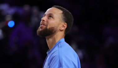 A man with a beard and short hair wearing a light blue long-sleeve shirt looks upward against a dark, blurry background.