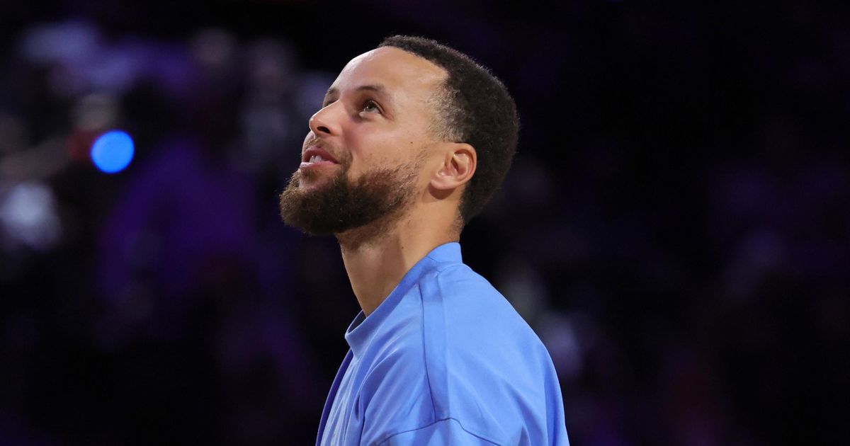 A man with a beard and short hair wearing a light blue long-sleeve shirt looks upward against a dark, blurry background.
