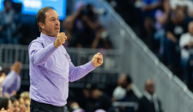 A man in a light purple shirt and black pants clenches his fists and appears to be shouting, standing in front of a cheering crowd.