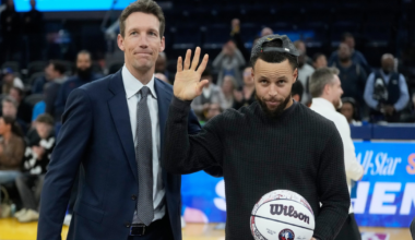 A man in a black sweater and backwards cap waves while holding a signed Wilson basketball, standing next to a man in a blue suit on a basketball court.