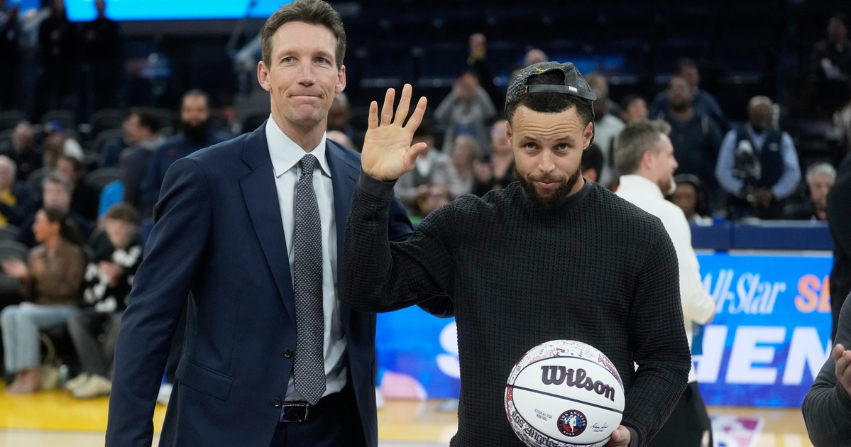 A man in a black sweater and backwards cap waves while holding a signed Wilson basketball, standing next to a man in a blue suit on a basketball court.