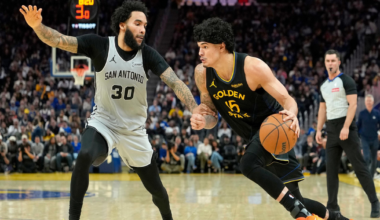 A Golden State player dribbles past a San Antonio defender in a closely contested basketball game with a referee in the background.