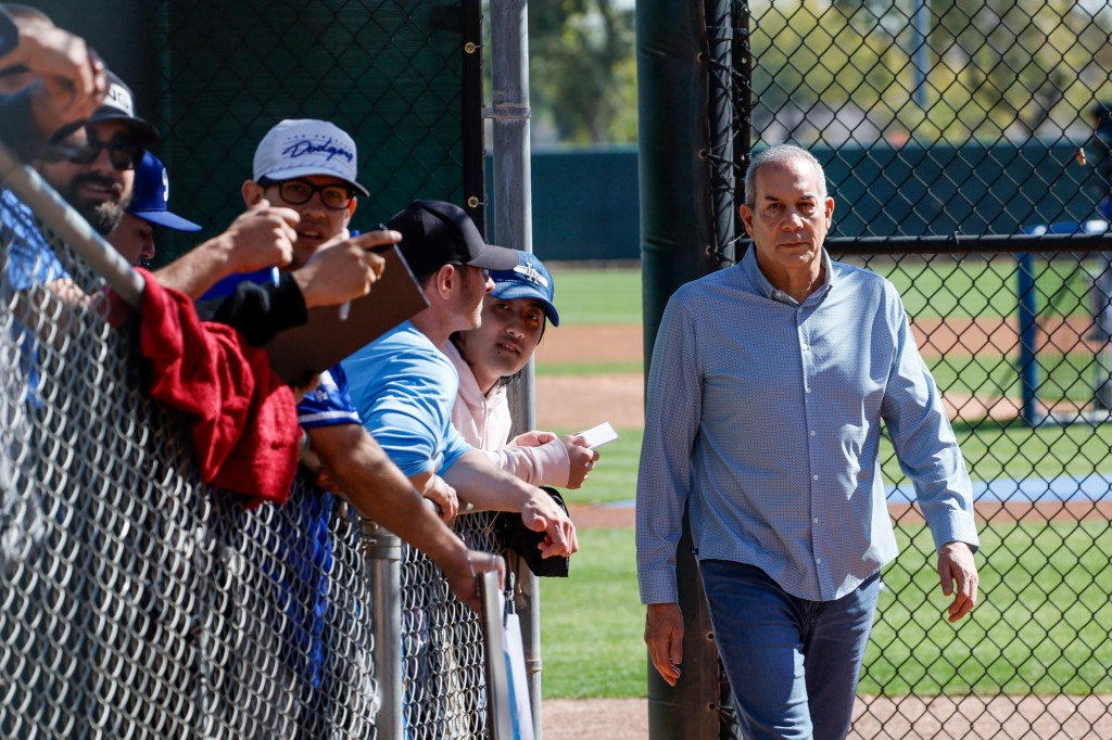 Fans at a baseball game lean over a chain-link fence, waiting for an autograph from a man in a light blue patterned shirt.