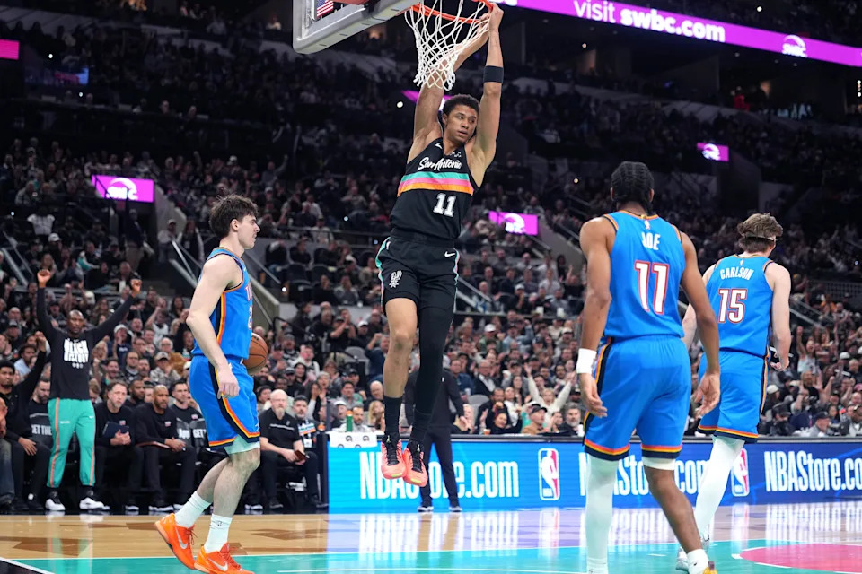 Feb 4, 2026; San Antonio, Texas, USA; San Antonio Spurs forward Carter Bryant (11) hangs on to the rim after dunking over Oklahoma City Thunder guard Brooks Barnhizer (23) during the first half at Frost Bank Center. Mandatory Credit: Scott Wachter-Imagn Images