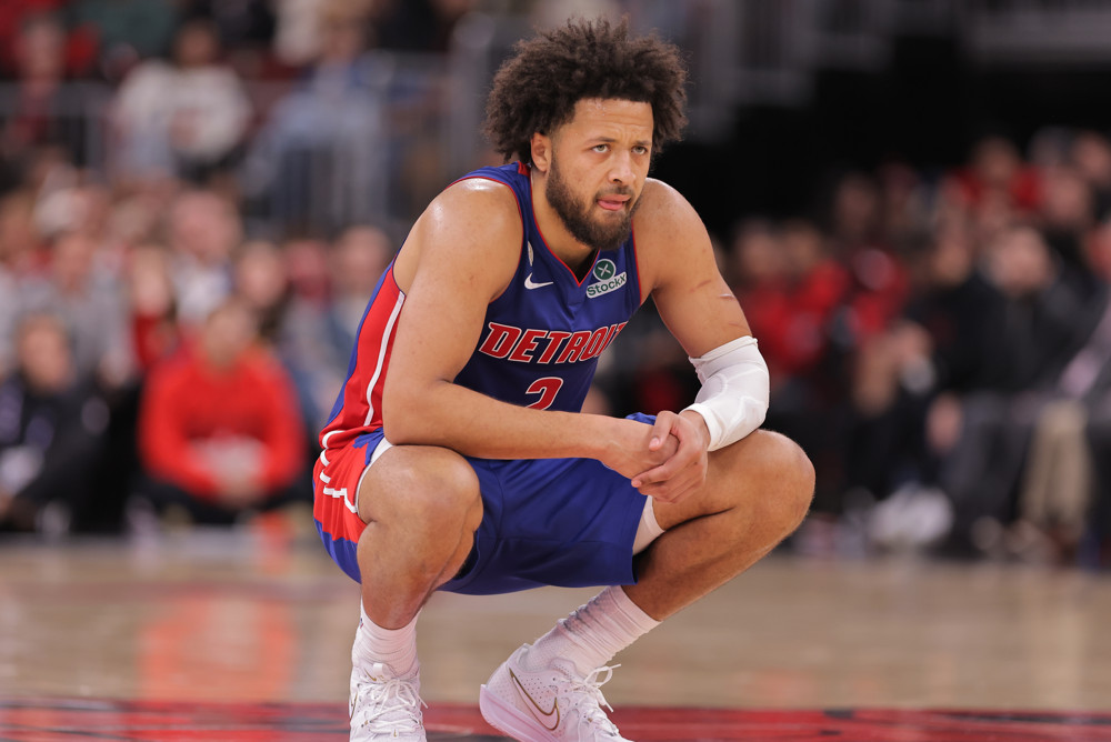 Cade Cunningham, wearing a blue Detroit Pistons jersey, an afro, and a beard, crouches on a basketball court as if in a baseball catcher's stance.