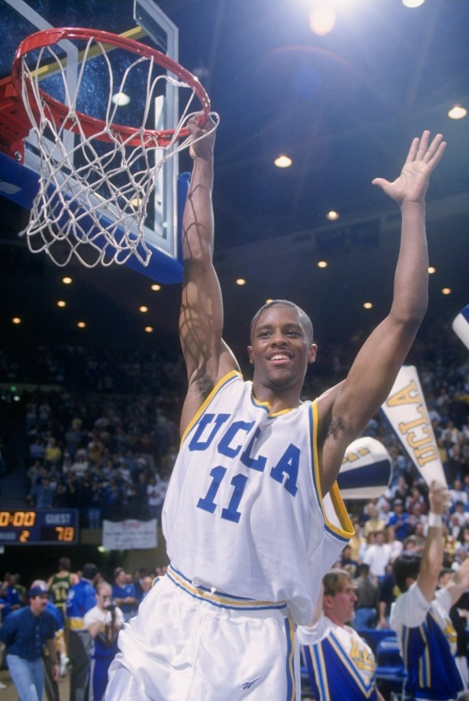 Tyus Edney of the UCLA Bruins celebrating by holding onto the basketball rim with one hand and raising his other arm, smiling.