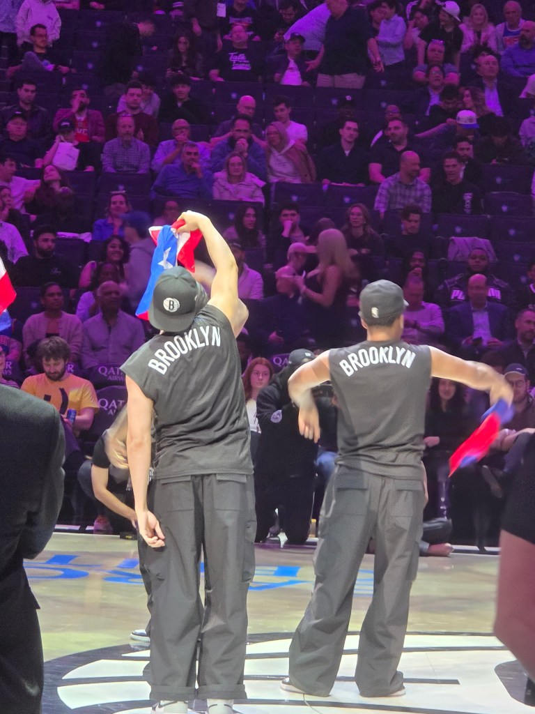 A Brooklynette and dancers with Team Hype wave Haitian flags during the West Indian Carnival-themed night at Barclays Center in Brooklyn on Tuesday, March 10, 2026. Photos by Darlie Gervais for The Haitian Times