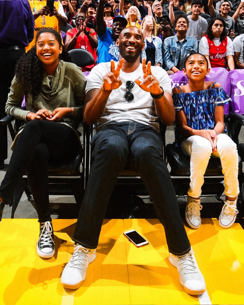 Kobe Bryant smiling and making a peace sign, sitting between his two daughters who are also smiling, in a stadium audience.