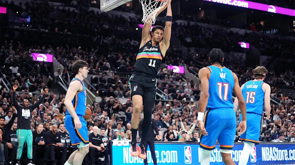 Feb 4, 2026; San Antonio, Texas, USA; San Antonio Spurs forward Carter Bryant (11) hangs on to the rim after dunking over Oklahoma City Thunder guard Brooks Barnhizer (23) during the first half at Frost Bank Center. Mandatory Credit: Scott Wachter-Imagn Images