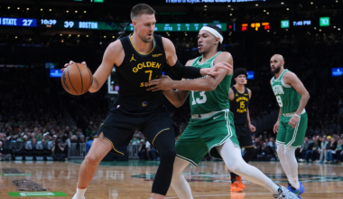 A Golden State player holds the basketball while defending against a Boston player in green during a lively basketball game.