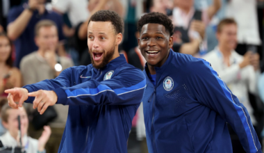 Two men in matching blue athletic jackets smile and gesture enthusiastically toward something off-camera, with a crowd blurred in the background.
