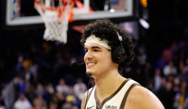 A basketball player wearing a white "The Town" jersey and headset smiles on the court with a basketball hoop and crowd blurred in the background.