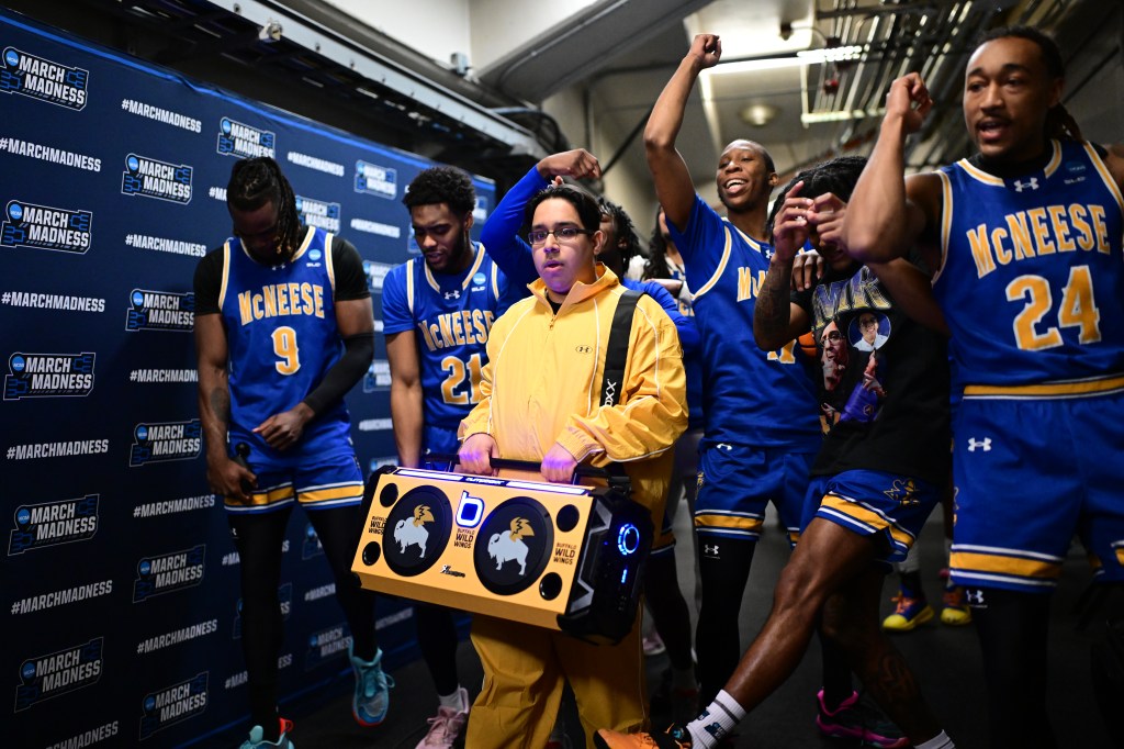 A student manager in a yellow tracksuit carrying a Buffalo Wild Wings boombox walks with McNeese State basketball players in blue and gold jerseys at the NCAA Men's Basketball Tournament.
