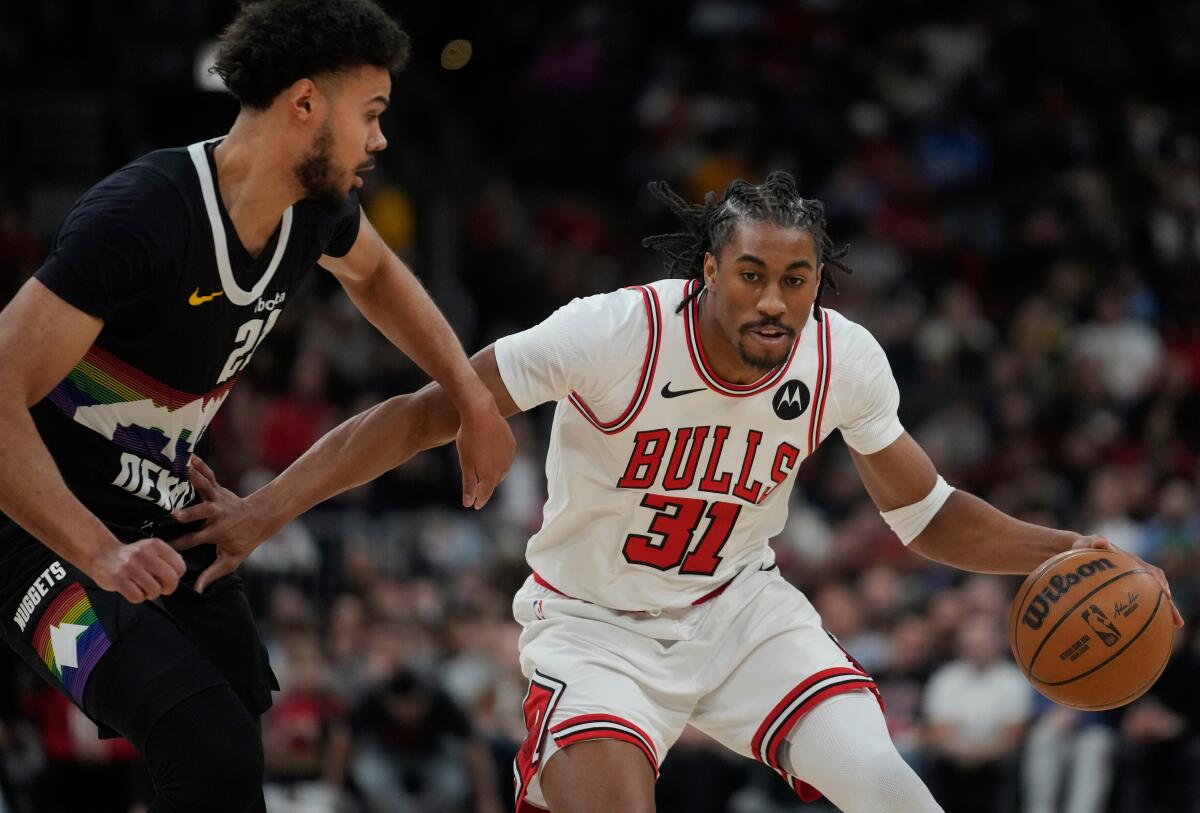 A man in a Chicago Bulls uniform bends down while dribbling the ball, using his right off to fend off a defender