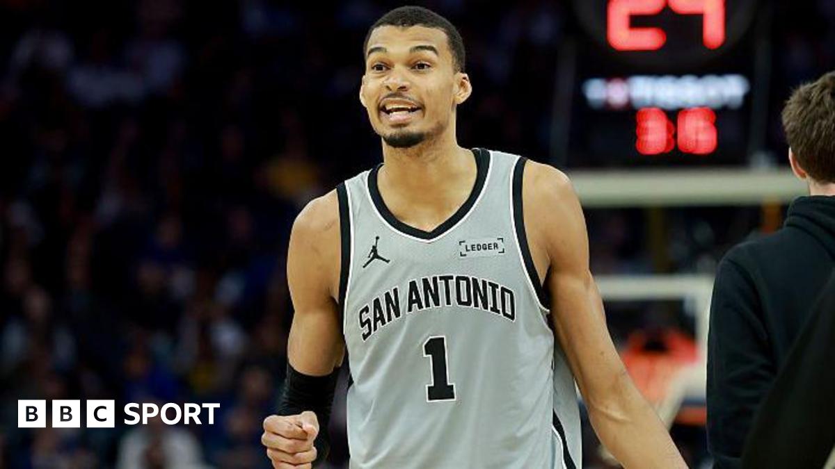Victor Wembanyama holds up his fist and is wearing a grey vest