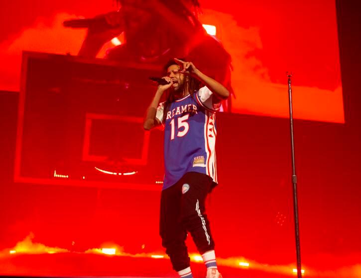 J. Cole holds a microphone to his mouth and points to the crowd while wearing a basketball jersey that says 'Dreamer 15'