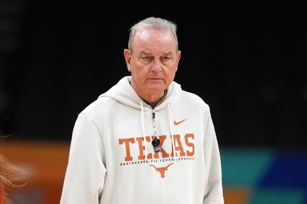 Vic Schaefer in a white "Texas" hoodie stands against a dark background. He has a serious expression and a whistle around his neck.