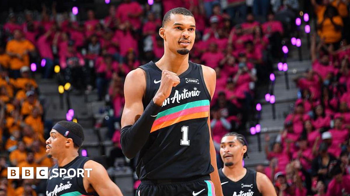 Victor Wembanyama, wearing a black San Antonio vest with a turquoise, pink, and orange stripe across the chest, punches his right fist in celebration and smiles