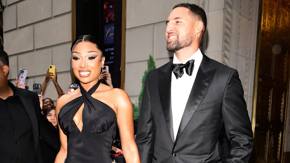 Megan Thee Stallion in a black dress and Klay Thompson in a tuxedo, smiling and walking outside a building.