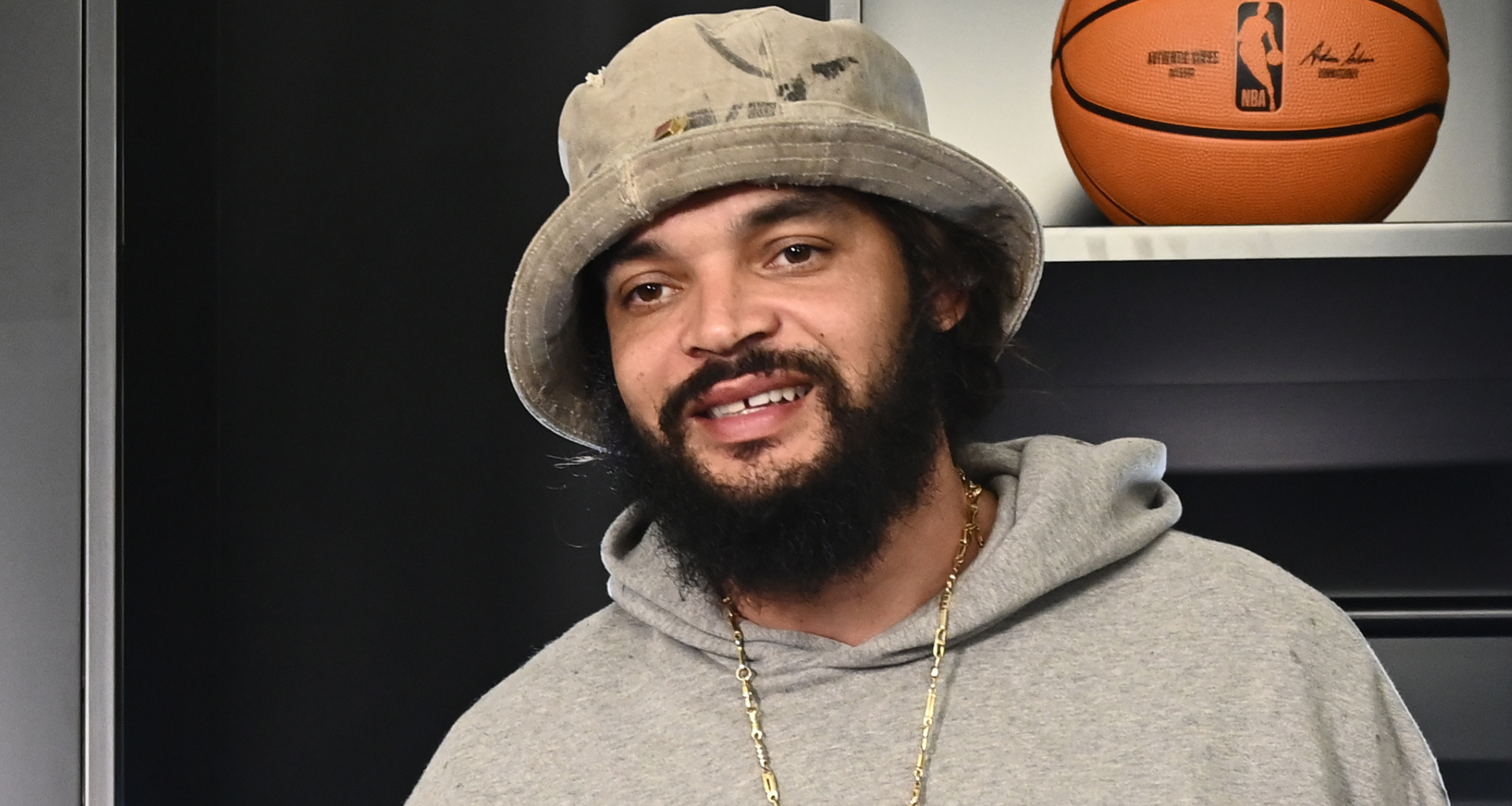 Joakim Noah, wearing a gray hoodie and a bucket hat, is standing indoors with a basketball in the background.