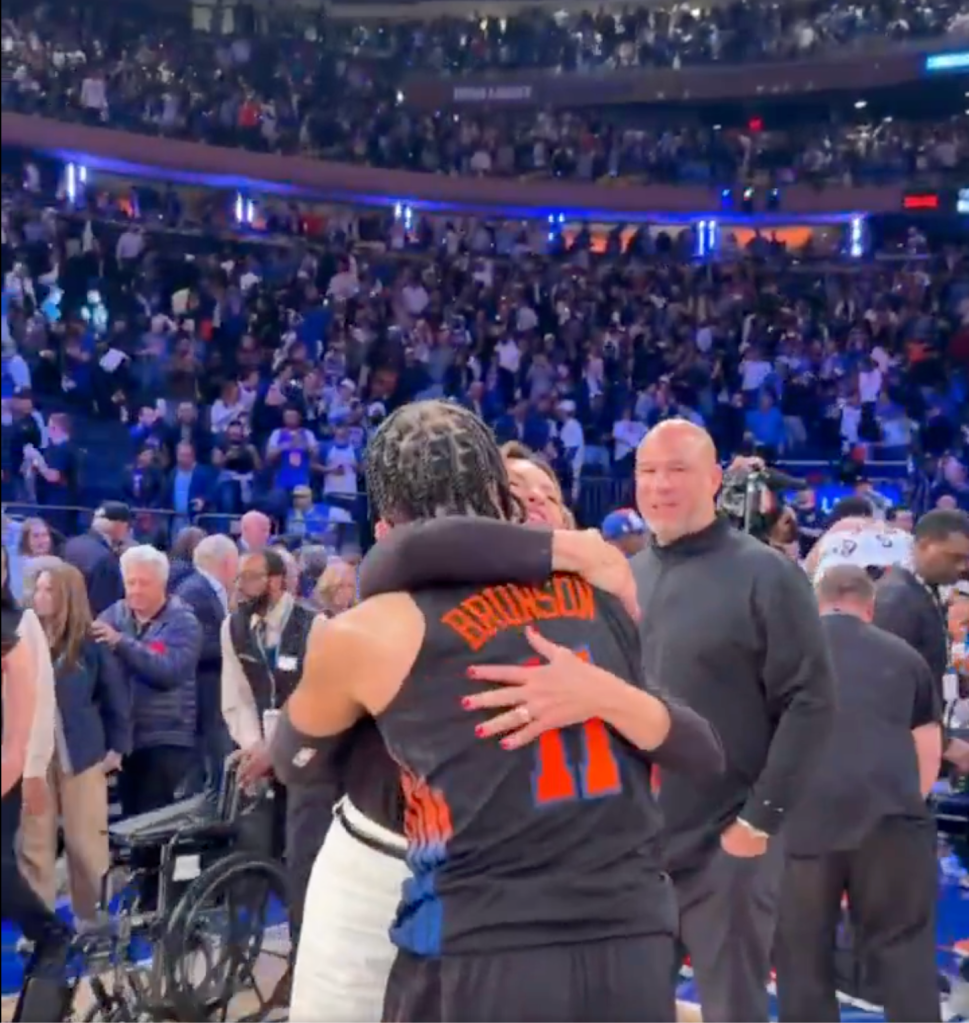 A person wearing a "Brunson 11" jersey hugging another person on a basketball court with a crowd in the background.
