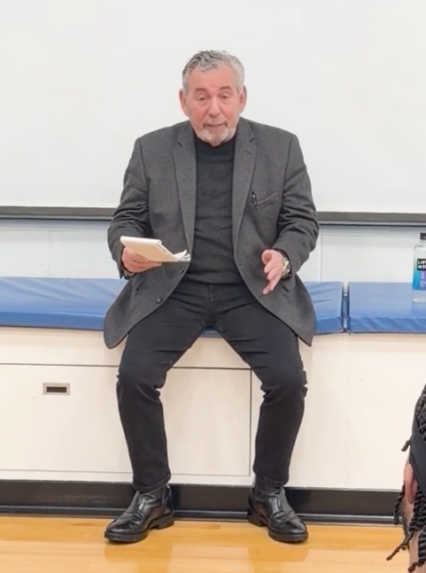 A man with gray hair and a beard, wearing a dark jacket and pants, sits on a bench while holding papers and gesturing.