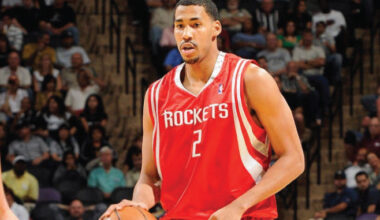 Basketball player in a red Houston Rockets jersey #2 dribbling the ball during a game with fans in the stands behind him.