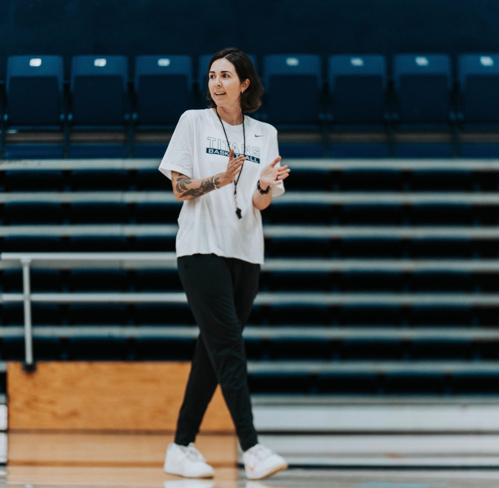 Cal State Fullerton coach Sammy Doucette paces the sidelines while encouraging her players during practice. The arena seating level is blurred in the background