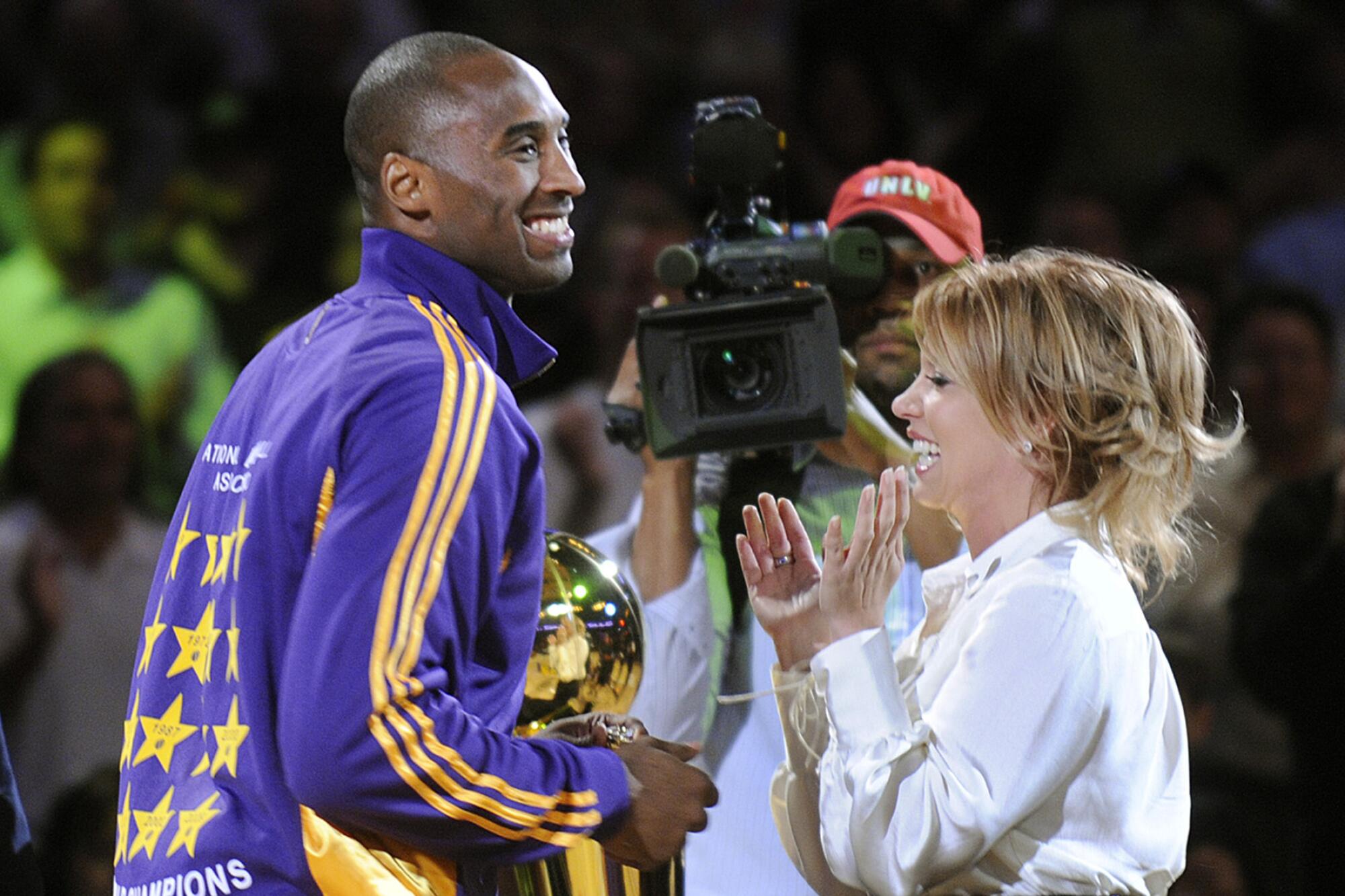 Lakers star Kobe Bryant and Lakers governor Jeanie Buss smile and stand beside each other on a basketball court. 
