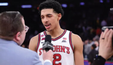 Basketball player in a St. John's jersey being interviewed on court, microphone held near his mouth, crowd in the background, jersey number 2 visible.
