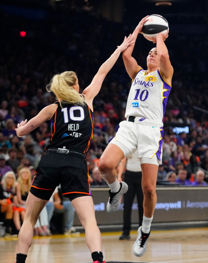 Kelsey Plum in a white and purple Sparks jersey shoots the ball as a player in a black and orange jersey defends.