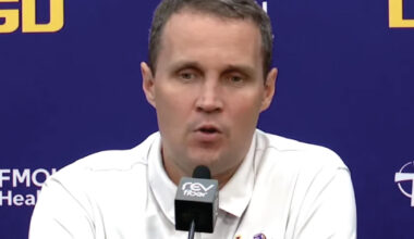 Man in a white polo shirt speaks into a microphone at a press conference with a blue backdrop and logos behind him.