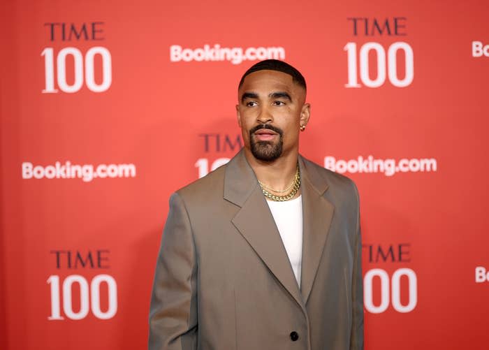 A person poses at a TIME 100 event, wearing a stylish beige blazer and chain necklace, with a branded backdrop in the background