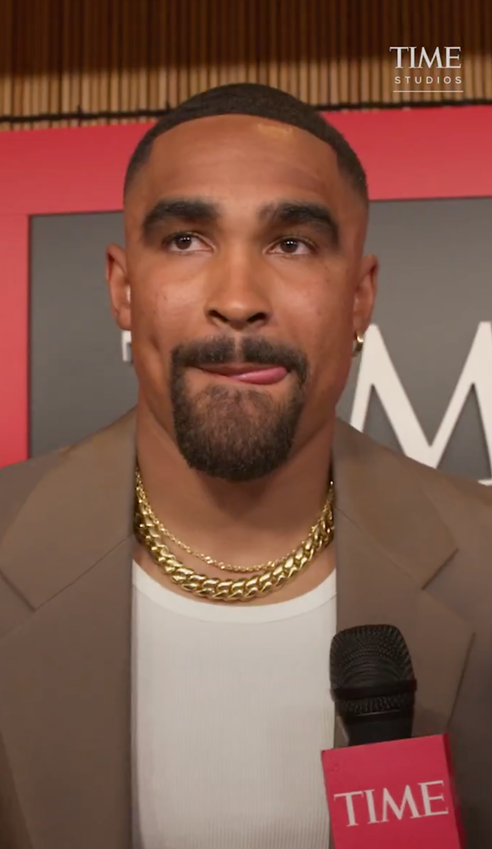 Person in suit with two chain necklaces, holding a microphone, standing in front of a backdrop with "TIME" partially visible