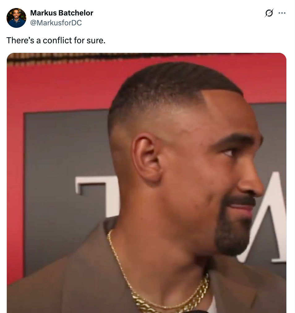 Man with close-cropped hair, wearing a suit and gold chain, stands against a backdrop, looking to the side