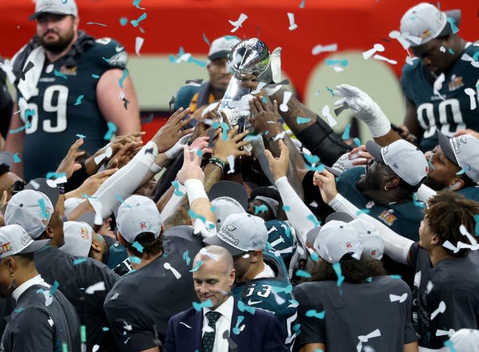 Team celebrates with trophy amid falling confetti on a sports field. Players in uniforms extend arms towards the trophy