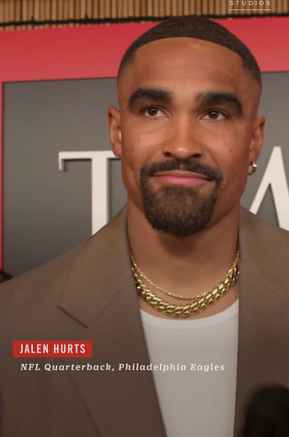 Person in a suit with gold chains at a formal event. The background includes a logo with overlapping letters, partially visible