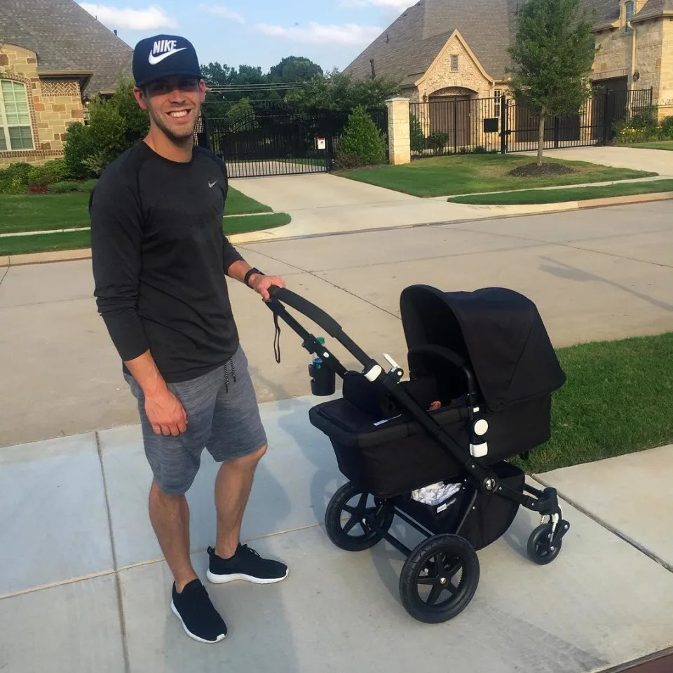 Justin Tucker wearing a black baseball hat, black shirt, and grey shorts, smiling while holding a baby stroller