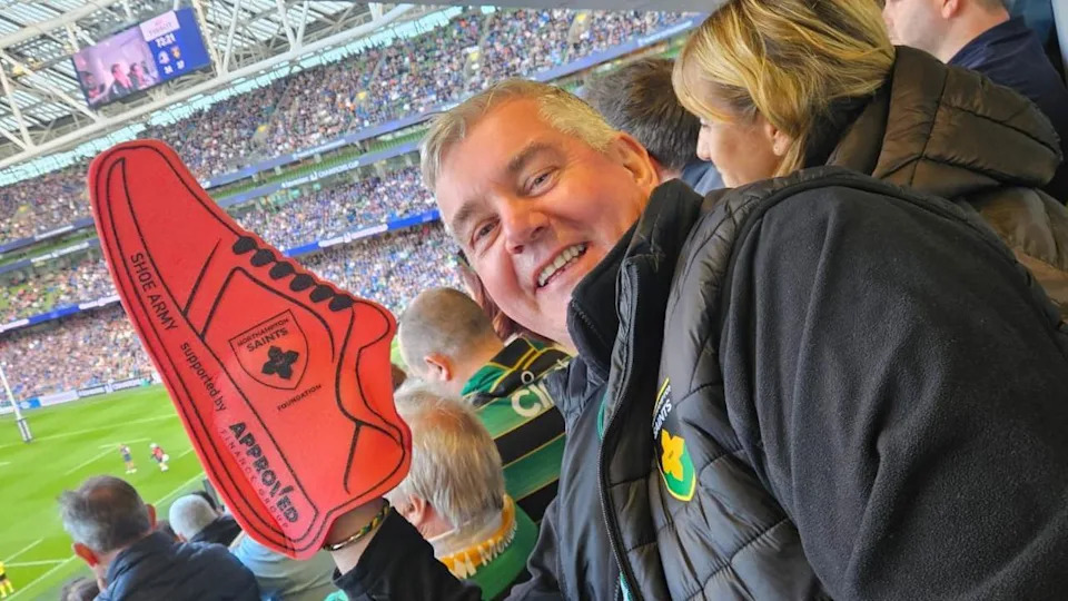 Colin Wells in a Northampton Saints jacket with a red foam shoe over his hand. He is standing against the backdrop of the Aviva Stadium in Dublin during the match against Leinster.