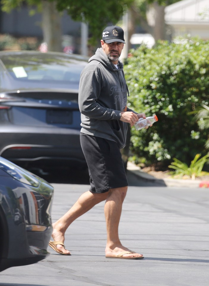 Aaron Rodgers wearing a baseball cap, gray hoodie, and black shorts while walking in a parking lot.