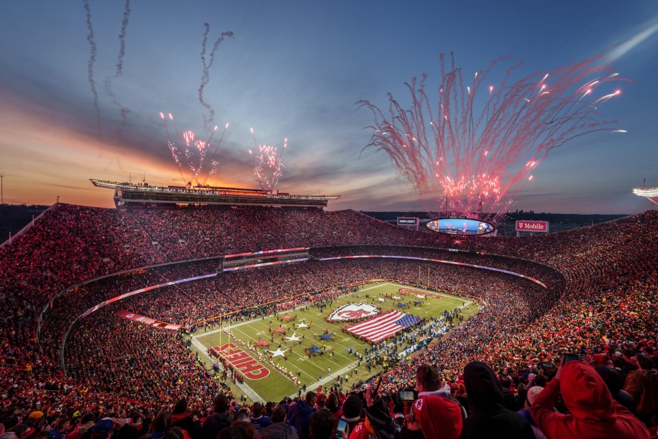 A full stadium at sunset with fireworks; an American flag and a large Chiefs logo are on the field.