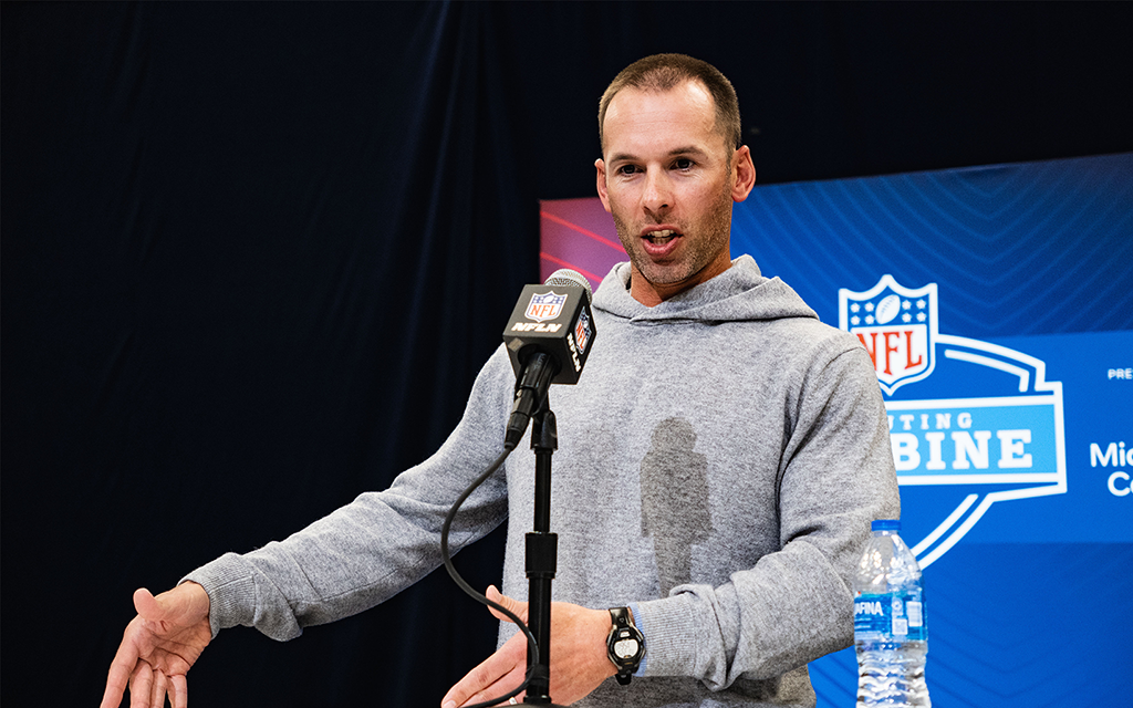 A person in a gray hoodie speaks into a microphone at an NFL event, with a backdrop featuring the NFL Scouting Combine logo.