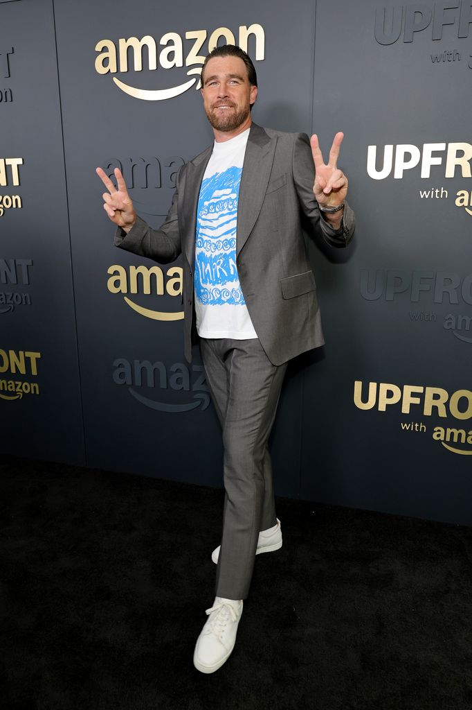 travis kelce in gray suit with slicked back hair throwing up peace signs