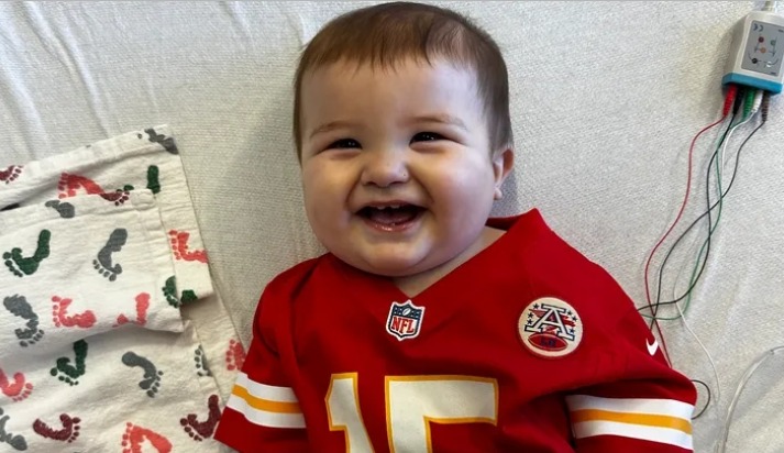 Smiling baby wearing a Kansas City Chiefs jersey in a hospital bed.