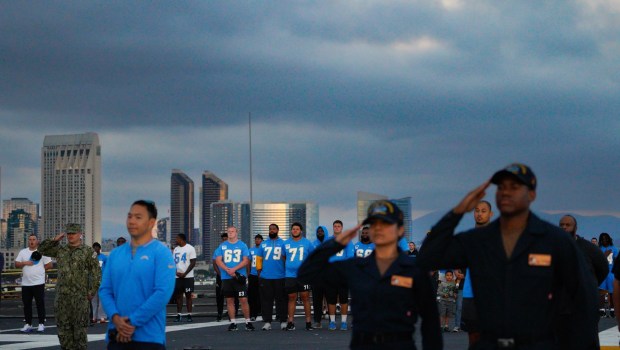 On the flight deck of the USS Abraham Lincoln (CVN 72) at NAS North Island in Coronado, CA, on Tuesday, July 22, 2025, sailors salute and the Los Angeles Chargers face the flag during the Evening Colors. The Navy and NFL come together to kick off the countdown of the big celebration the city will have in late Oct. when the Navy and the Marines celebrate their 250th anniversary. (Nelvin C. Cepeda / The San Diego Union-Tribune)
