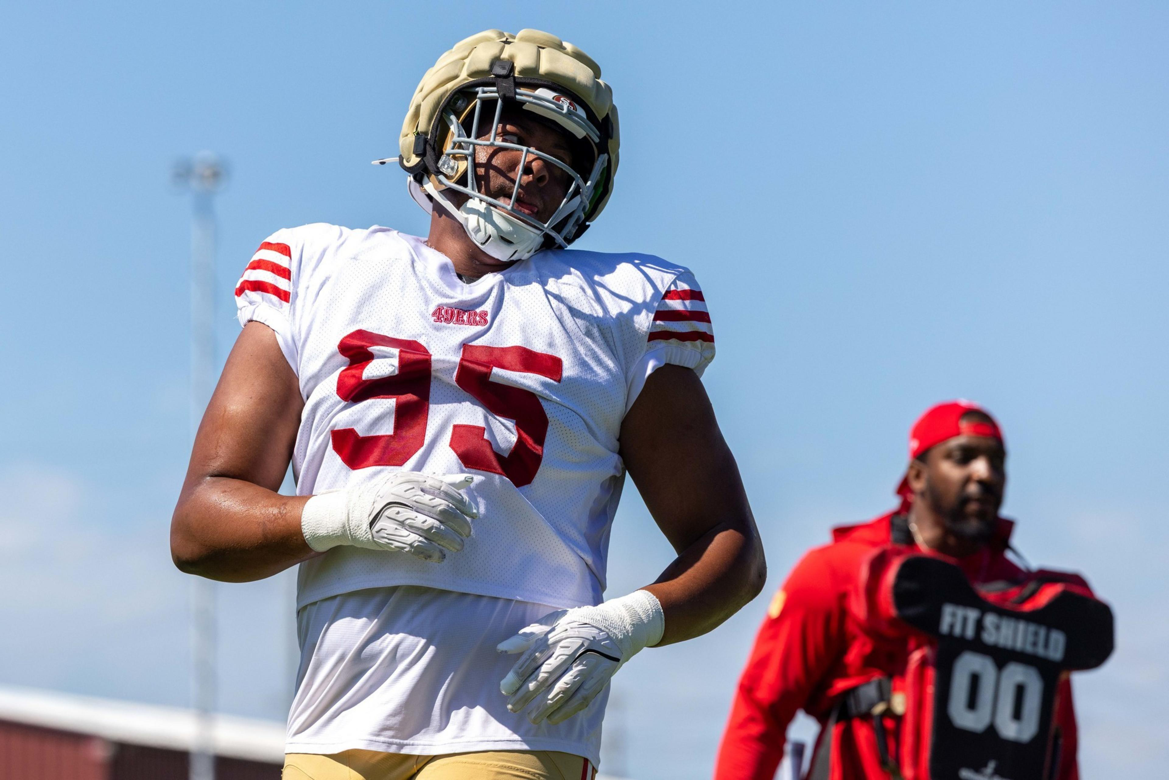 A football player wearing a white jersey with red number 95 and a gold helmet stretches while a coach dressed in red stands behind him.
