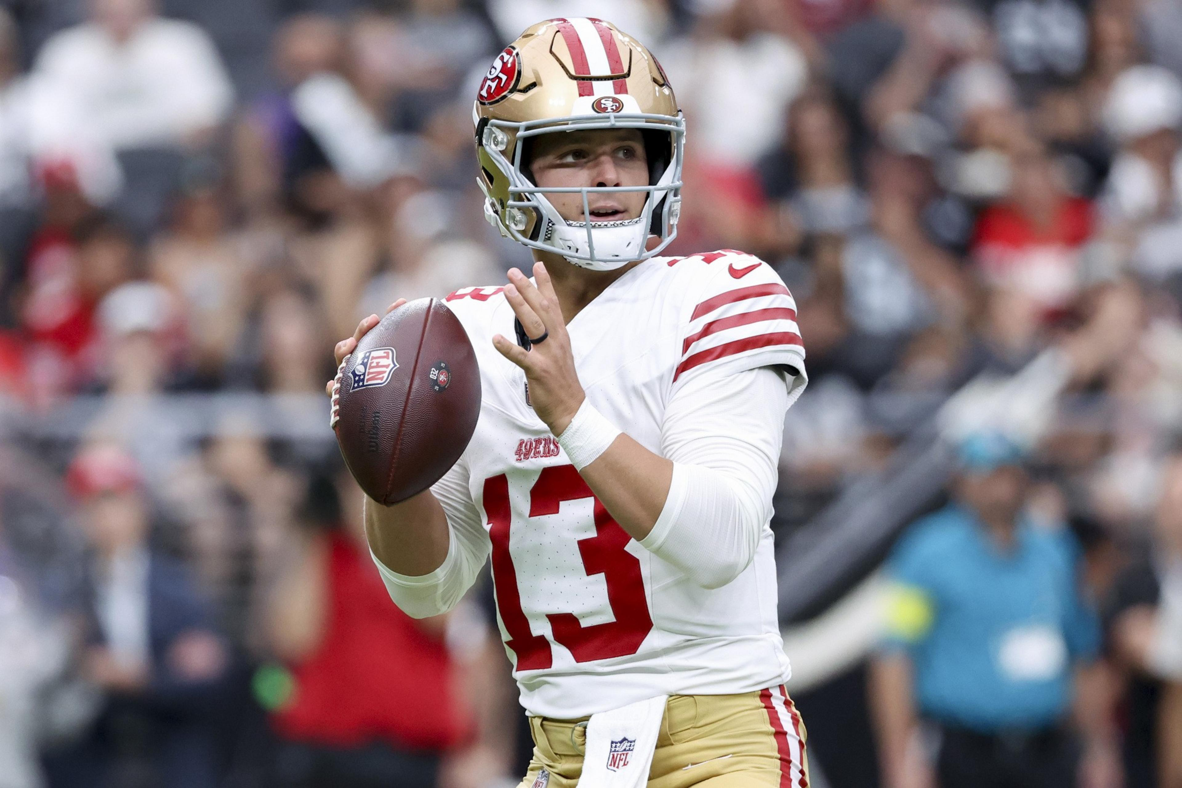 A football player in a white and gold 49ers uniform, number 13, prepares to throw a pass during a game with a blurred crowd in the background.