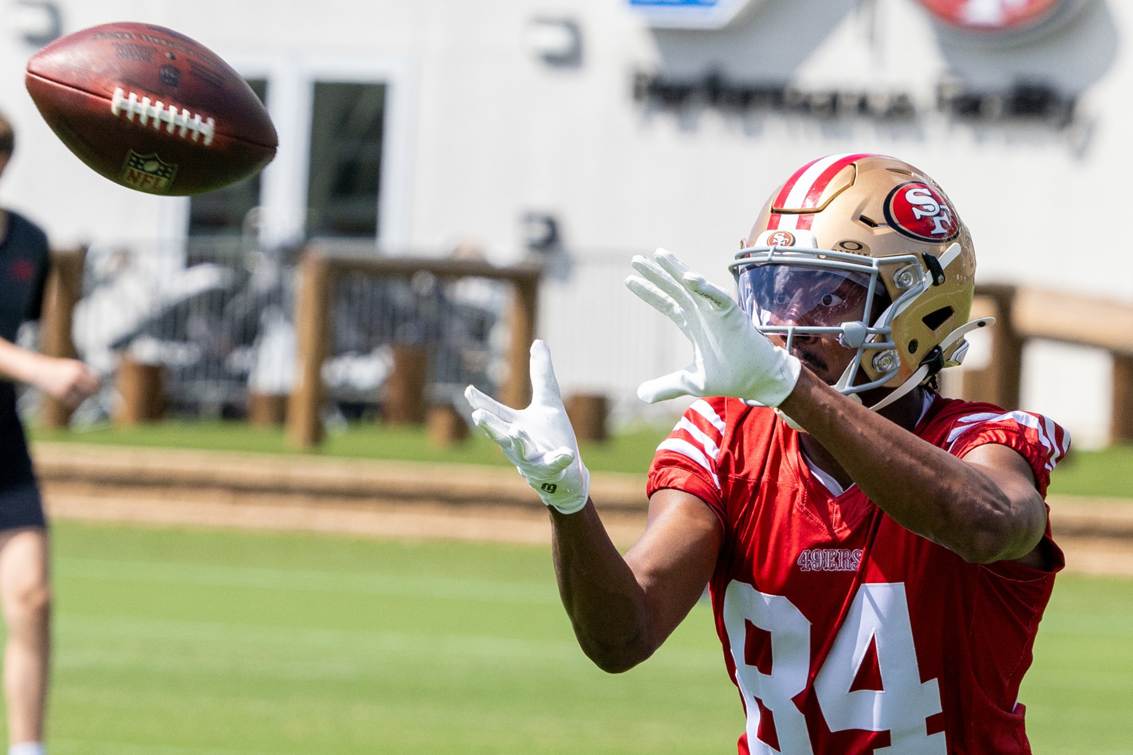 A football player wearing a red jersey and gold helmet is reaching out to catch a football. The background is a grassy field with blurred elements.