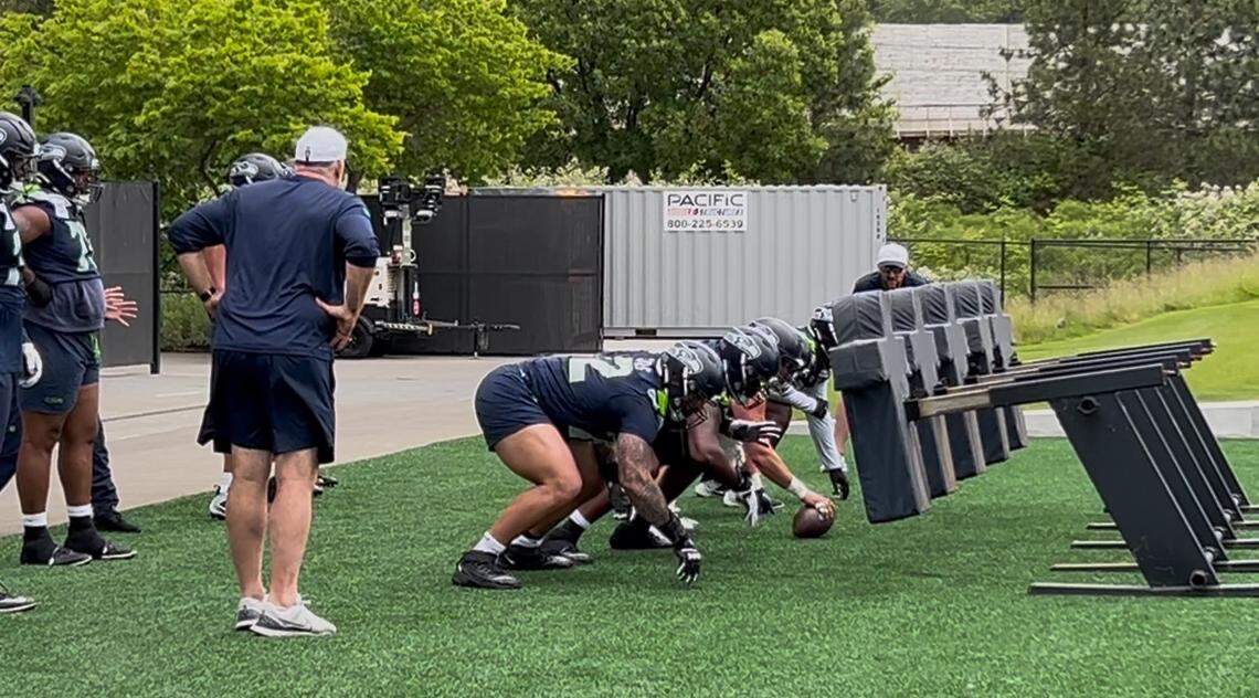 New Seahawks line coach John Benton (left in white cap), a veteran of 19 NFL seasons coaching, works right tackle Abe Lucas (72, center foreground) and the starting offensive line through a sled drill during organized team activities (OTAs) offseason practices June 4, 2025, at the Virginia Mason Athletic Center in Renton.