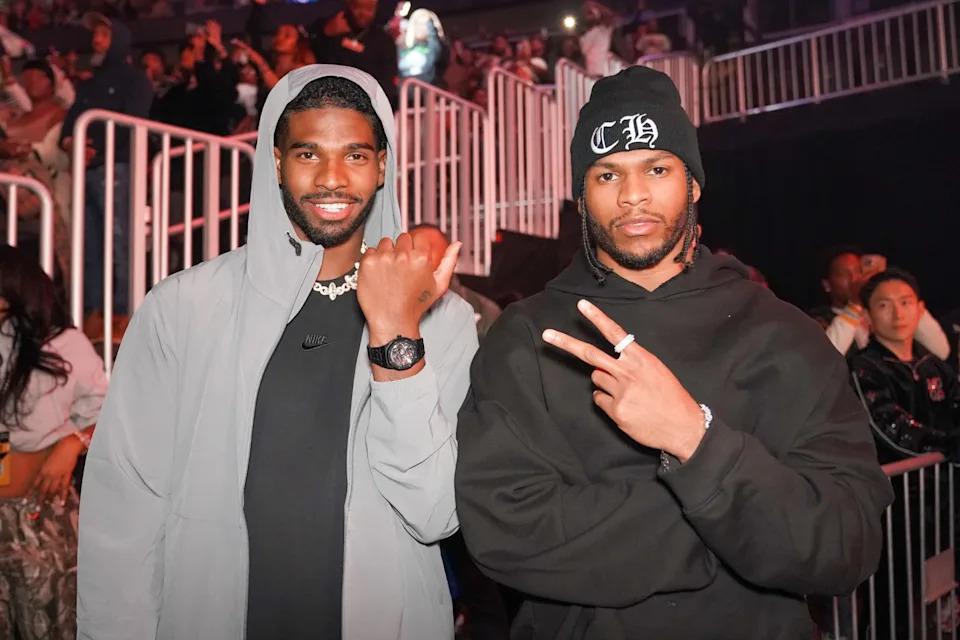 Shedeur and Shilo Sanders posing at AT&T Playoff Playlist Live!, one wearing a light jacket and watch, the other in a black hoodie and beanie, flashing peace signs.